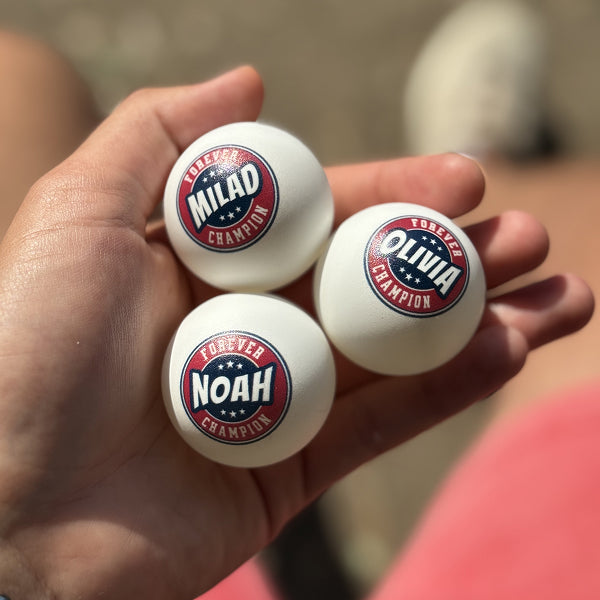 Three personalized ping pong balls with the names Olivia, Noah, and Milad held in a hand against a blurred background